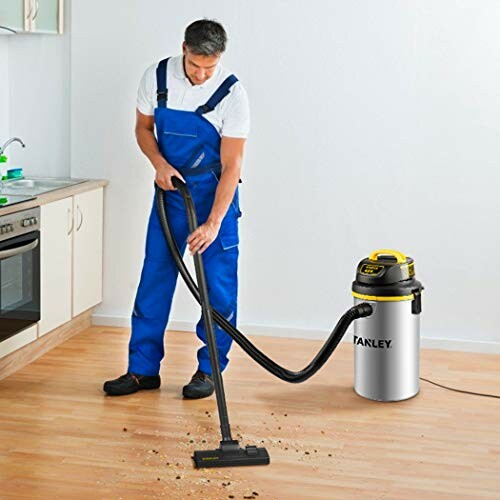 Man in blue overalls using a shop vacuum cleaner on a wooden floor.